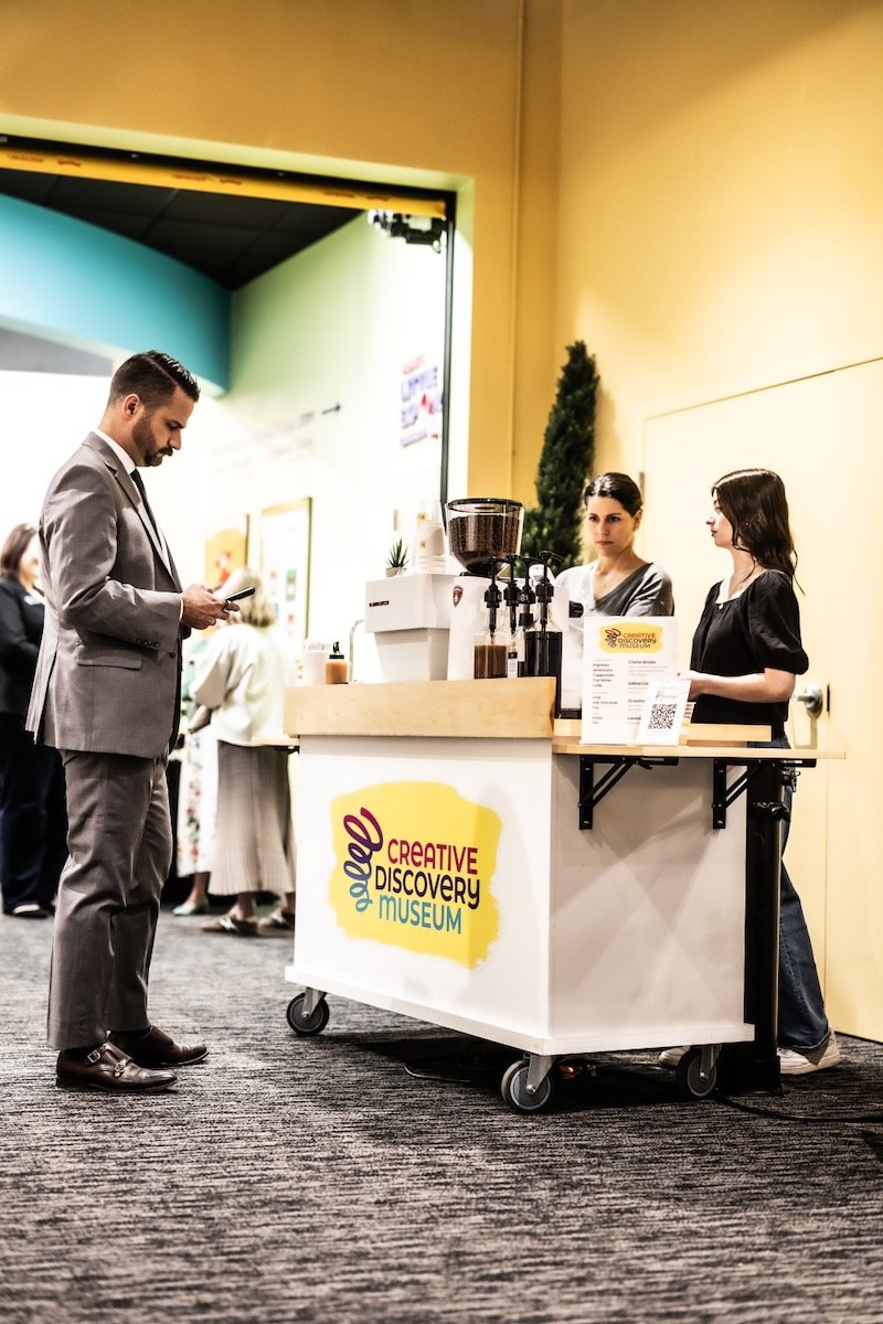 A man in a gray suit ordering coffee from two women behind a mobile coffee cart at the Creative Discovery Museum. The cart has a sign with the museum's logo.