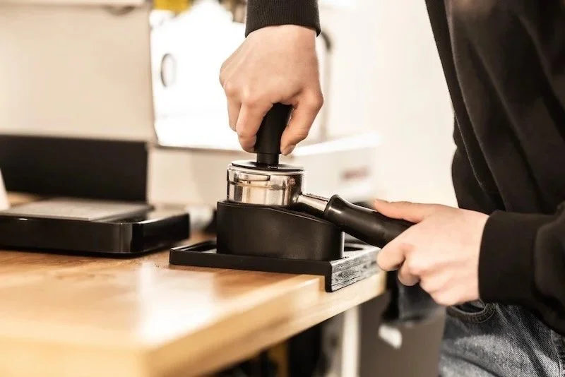 A person's hand pressing a tamper into coffee grounds in a portafilter during espresso preparation.