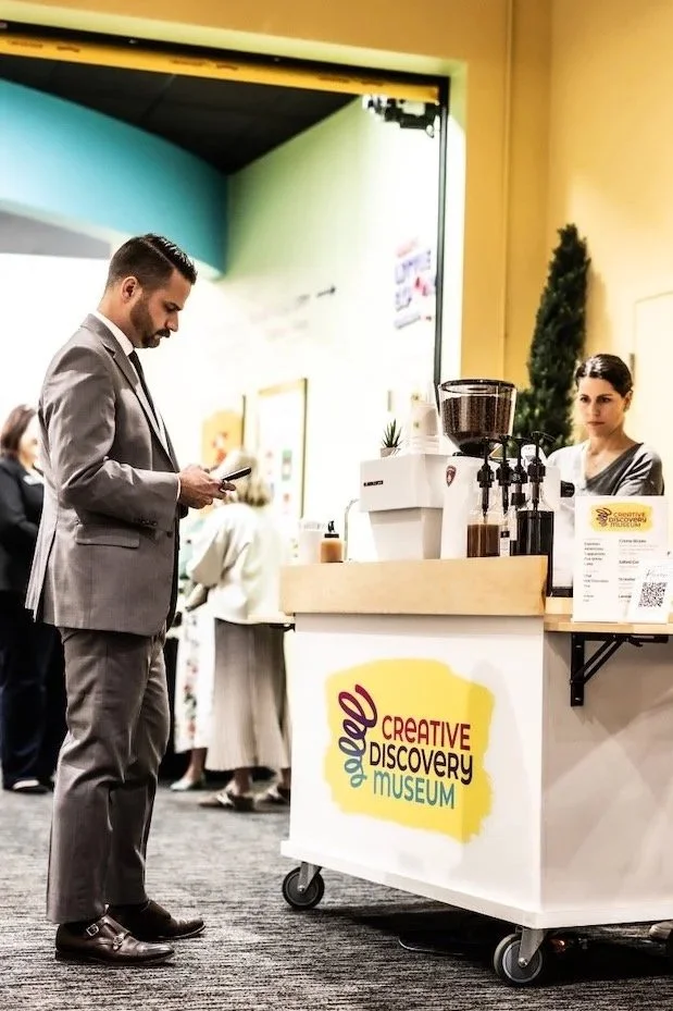 A man in a gray suit stands at a coffee cart with a banner that reads "Creative Discovery Museum". Two women behind the cart are preparing coffee. The setting appears to be indoors at an event or museum entrance.