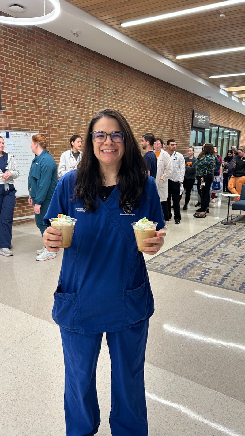 A woman wearing blue medical scrubs smiling and holding two cups coffee at rthe College of Veterinary Medicine at the University of Tennessee Institute of Agriculture with people in the background. She's happy about the quick and friendly service.