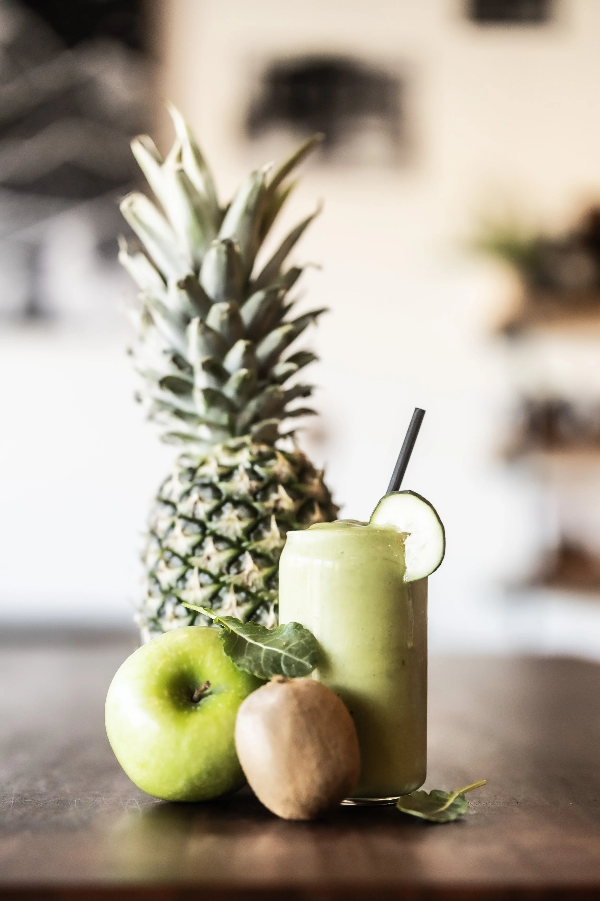 A pineapple, green apple, kiwi, and a green smoothie with a cucumber slice garnish on a wooden surface.