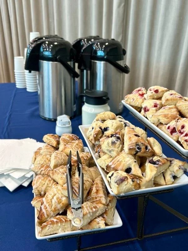Tray of assorted pastries including croissants and fruit-filled danishes, with coffee urns and cups in the background at a buffet table.