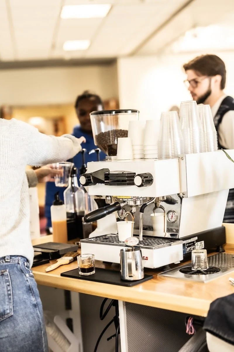 A barista prepares espresso drinks on a coffee machine in a café, with stacks of clear plastic cups and coffee beans in a container visible on the counter.