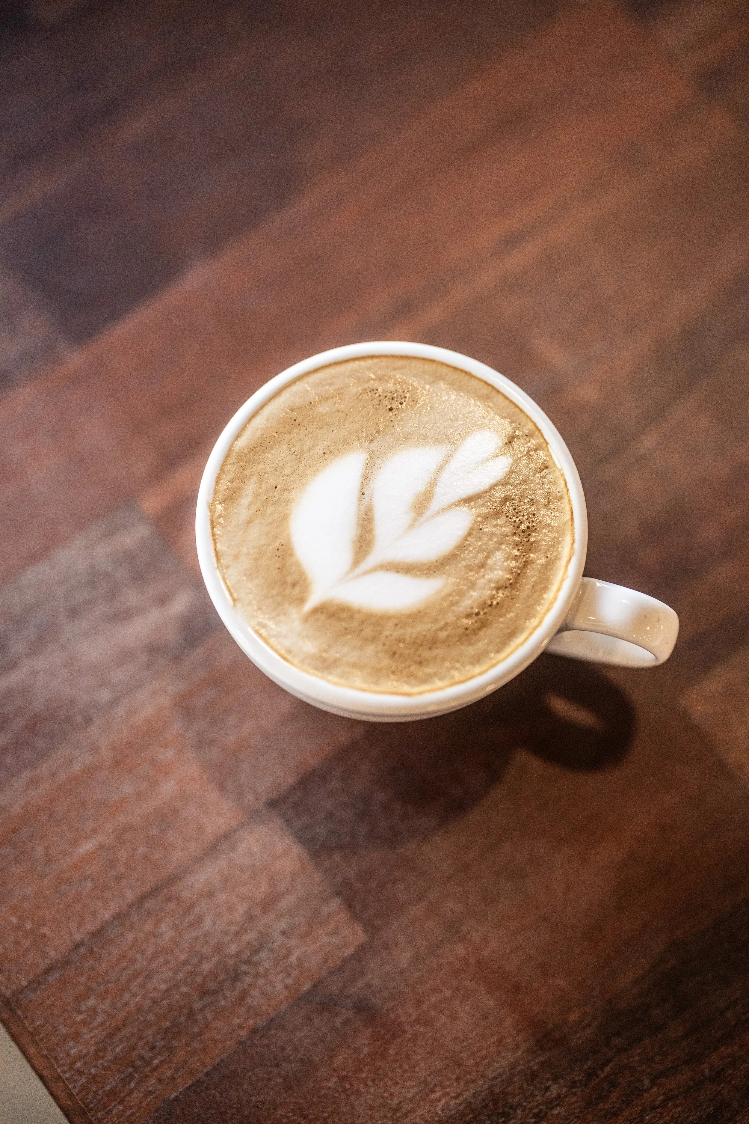 Top-down view of a latte with a leaf foam art design in a white mug on a wooden table.