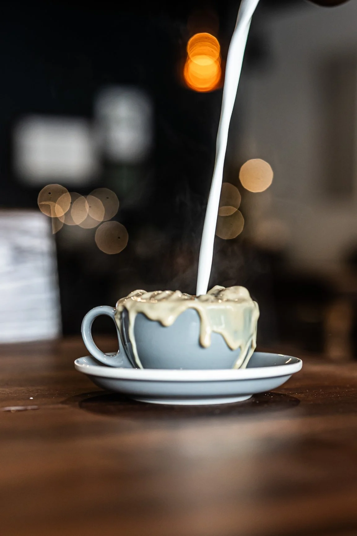 A white tea cup with a saucer underneath it, filled with a hot beverage, with cream or milk being poured into it, creating a splash on top. The background is blurred and features warm bokeh lights.