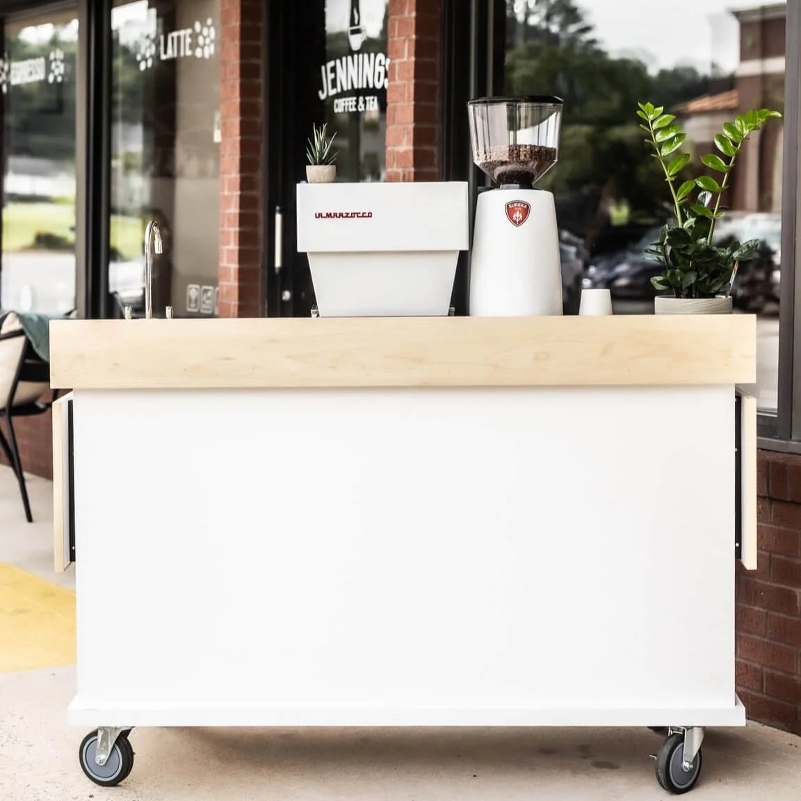 Mobile coffee cart with coffee grinder and potted plants outside a cafe.