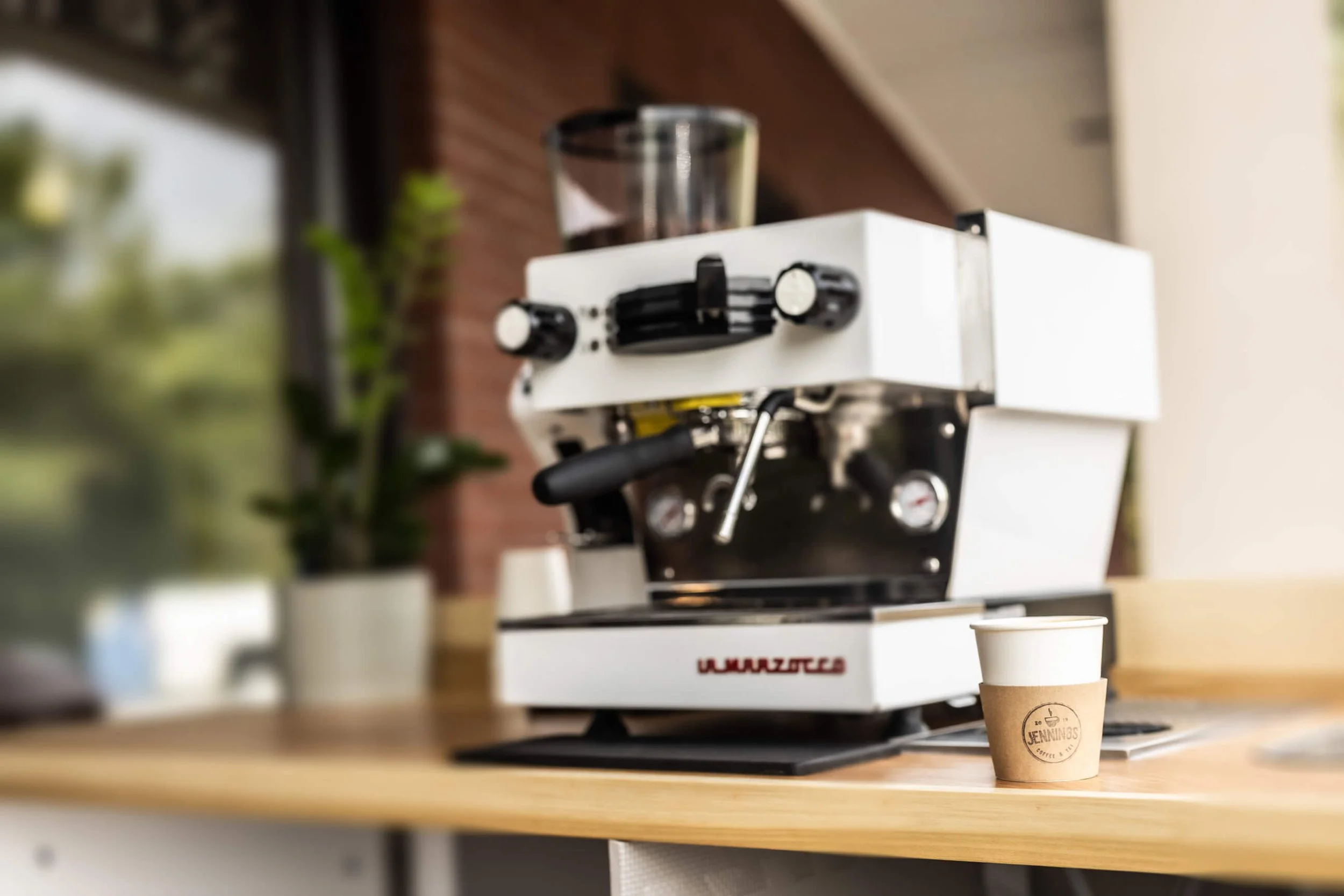 A white espresso machine on a wooden counter with a small paper cup in front of it