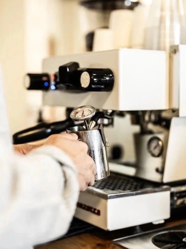 Close-up of a barista hand steaming milk in a metal pitcher using an espresso machine in a coffee shop.