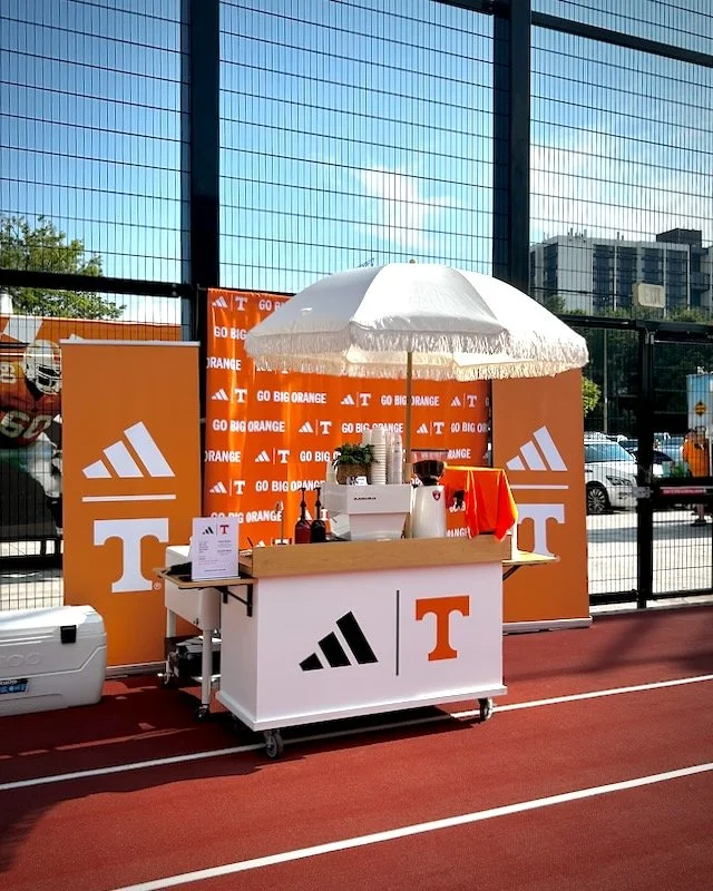 University of Tennessee Knoxville promotional event with orange and white banner reading 'GO BIG ORANGE', featuring the Tennessee and Adidas logos, set up on Dick's House of Sport track field, shaded by a white umbrella. Catered by The Coffee Cart.