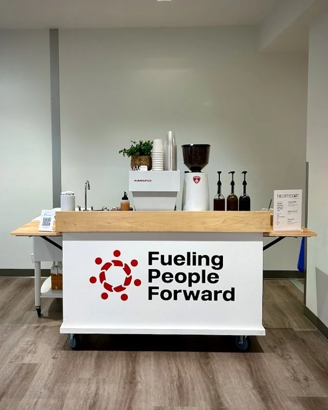 A coffee stand with a wooden top and a white front panel displaying a logo and the phrase "Fueling People Forward." The stand has a coffee grinder, cups, and bottles of syrup on top, with a small potted plant and a QR code on the side.