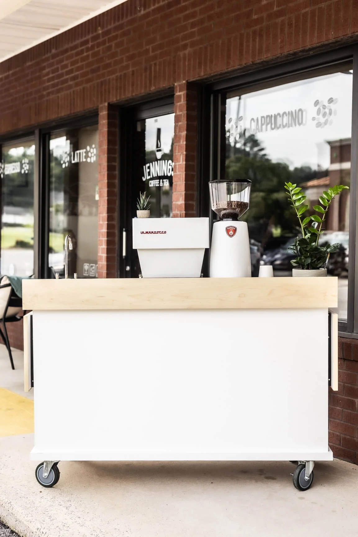 Coffee cart with a coffee grinder, plants, and a white container outside of a cafe.