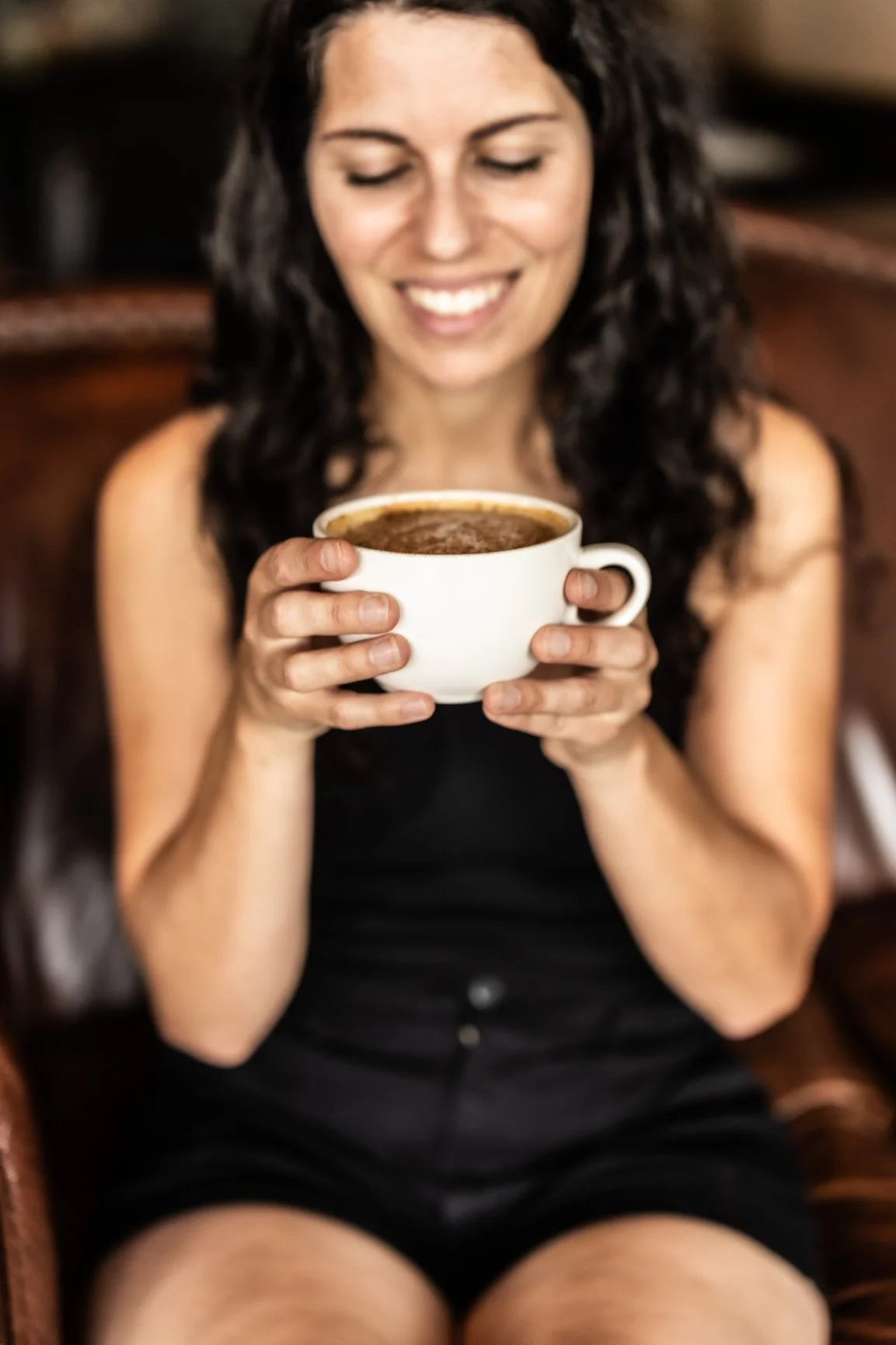 A woman with dark, curly hair smiling and holding a large cup of coffee or hot chocolate while sitting on a chair.