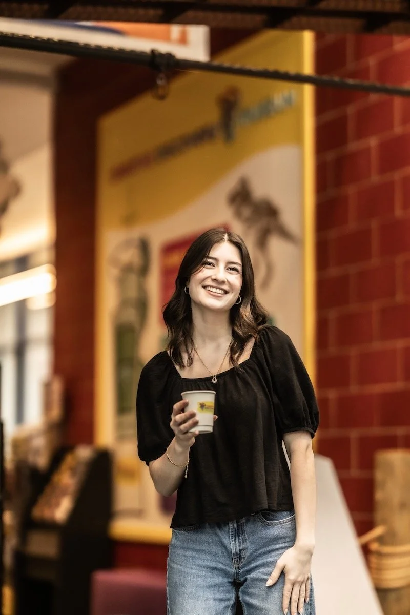 A young woman with dark hair in a black top and jeans holding a custom branded cup, smiling in the Creative Discovery Museum in Chattanooga  with warm lighting and colorful wall art.