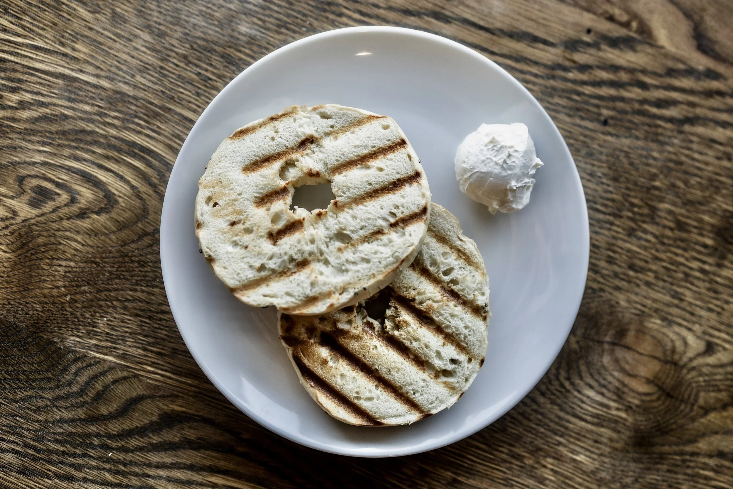 Two toasted bagel halves with grill marks on a white plate, served with a dollop of cream cheese, on a wooden table.