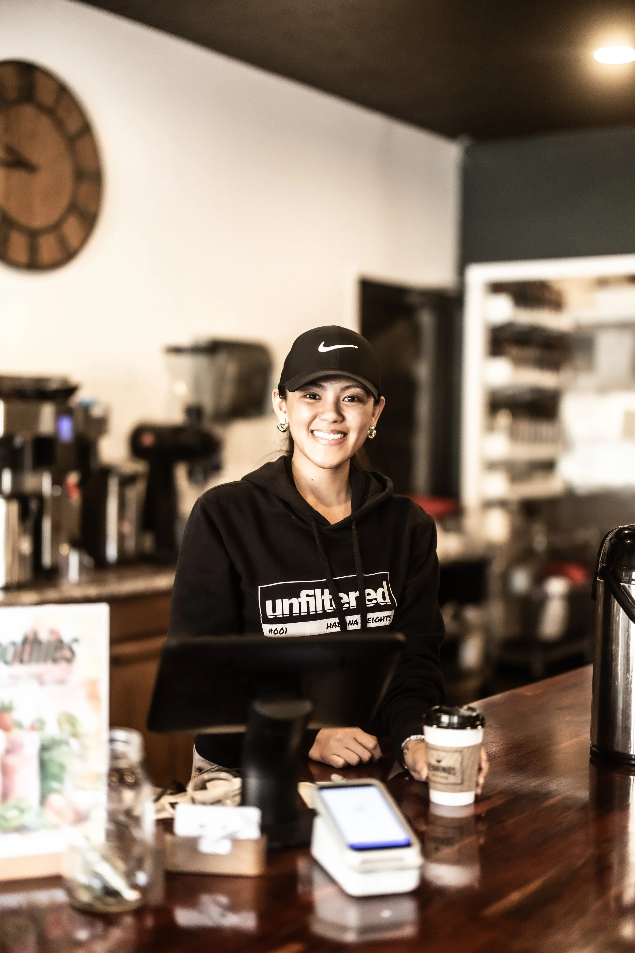 A smiling young woman with earrings, wearing a black hoodie with the word "uninspired" printed on it, and a black Nike cap, working at a coffee shop counter with coffee equipment and a disposable coffee cup.