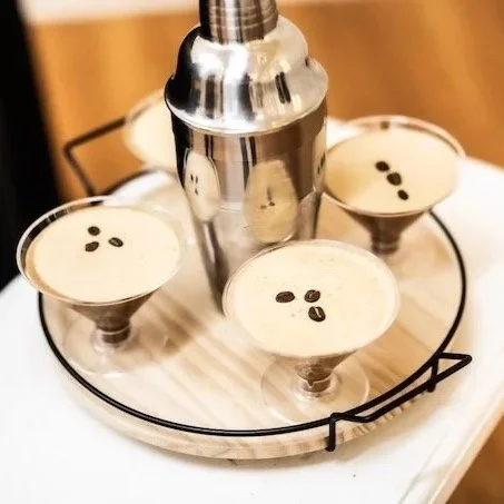 Four glasses of creamy espresso martinis topped with coffee beans on a round wooden tray, with a stainless steel whipped cream dispenser in the center.