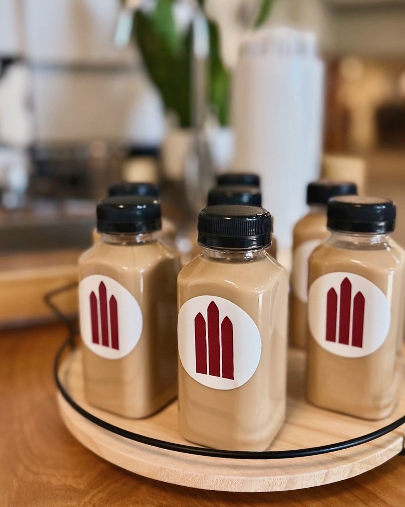 Bottles of latte or spread on a round wooden tray, with a logo featuring three vertical maroon shapes on a white background.
