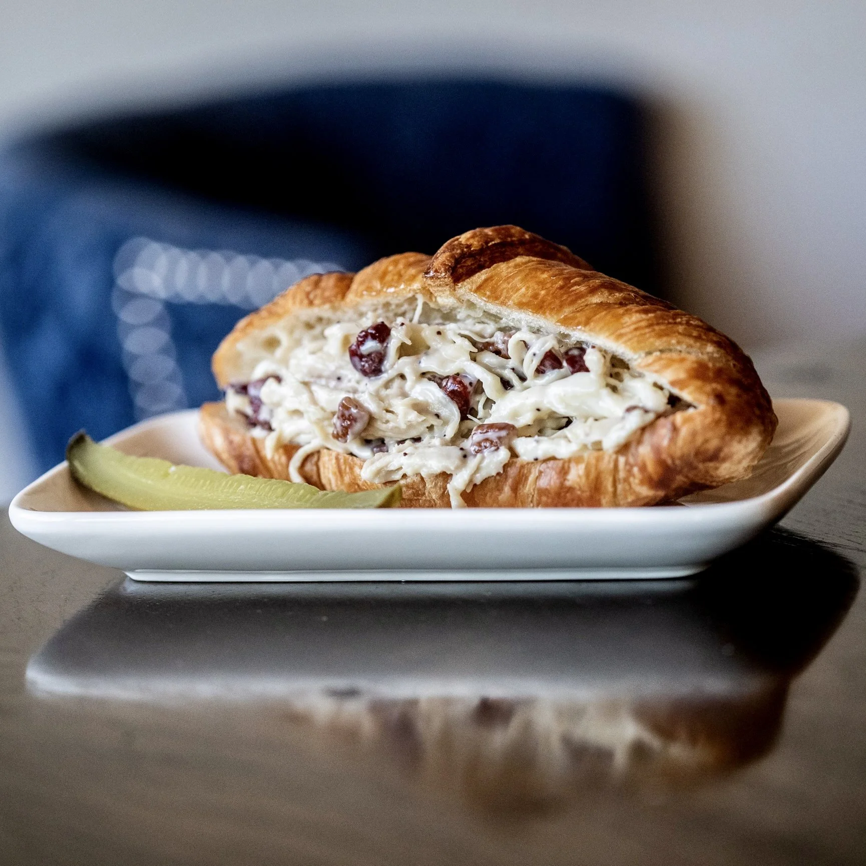 A croissant sandwich filled with creamy chicken salad containing cranberries, served on a white rectangular plate with pickle slices, on a dark surface with a blurred background.