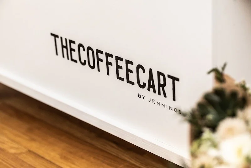 Sign reading "THECOFFEECART BY JENNINGS" on a white wall with wooden floor in the foreground and blurred flowers on the right.