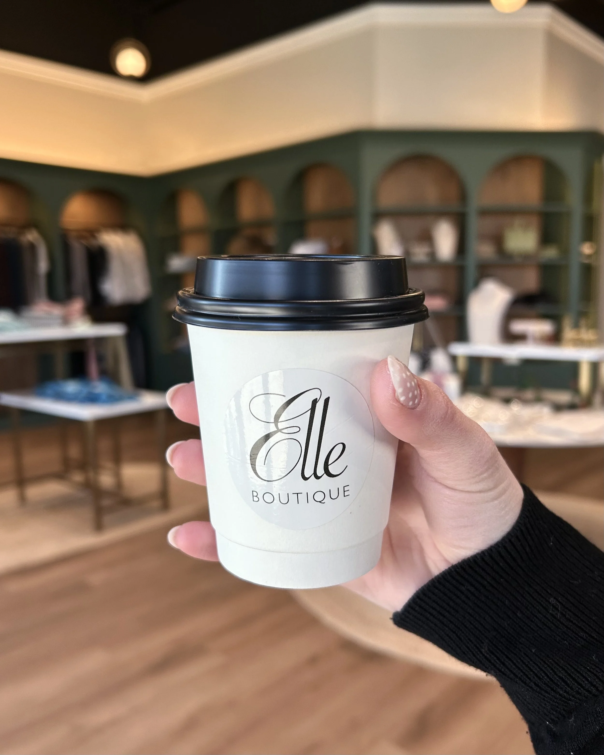Hand holding a white paper coffee cup with a black lid, labeled with custom branding, inside a boutique store with display shelves and clothing in the background.