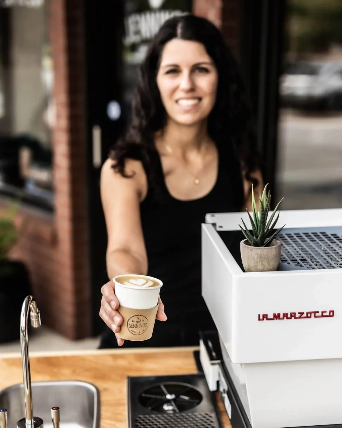 Woman smiling and handing a cup of coffee with latte art to the camera in an outdoor cafe setting.