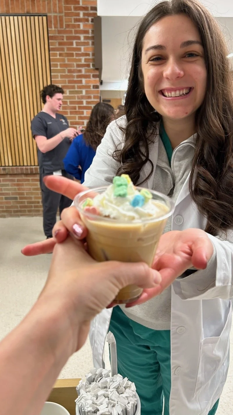 Smiling woman in medical scrubs receiving coffe with colorful toppings from a mobile event coffee bar barista. In the background, a man and woman stand near a brick wall.