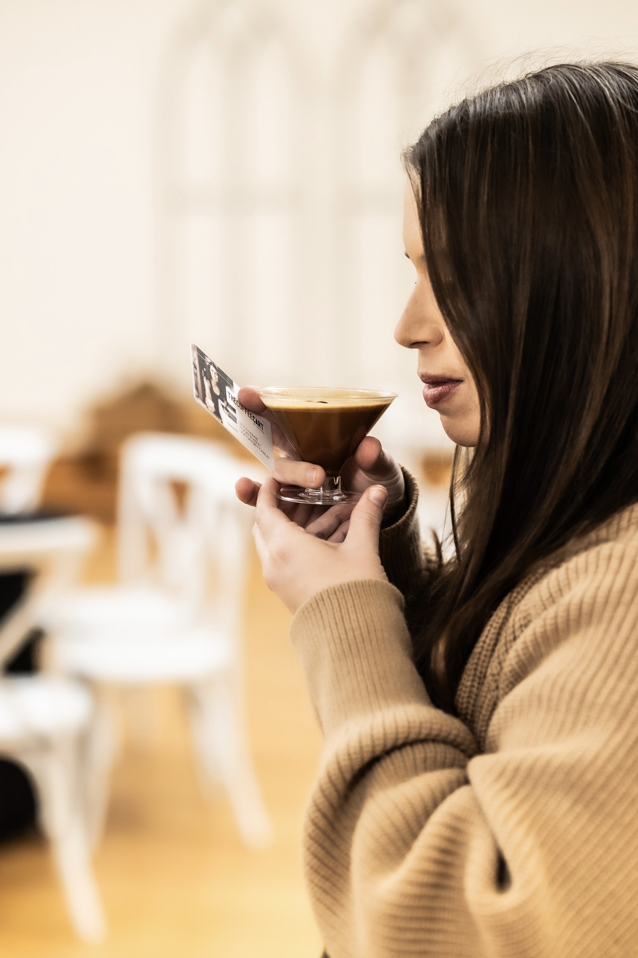 A woman with long dark hair holding a cocktail glass close to her face with one hand, and a ticket in the other, inside a well-lit room with wooden furniture.