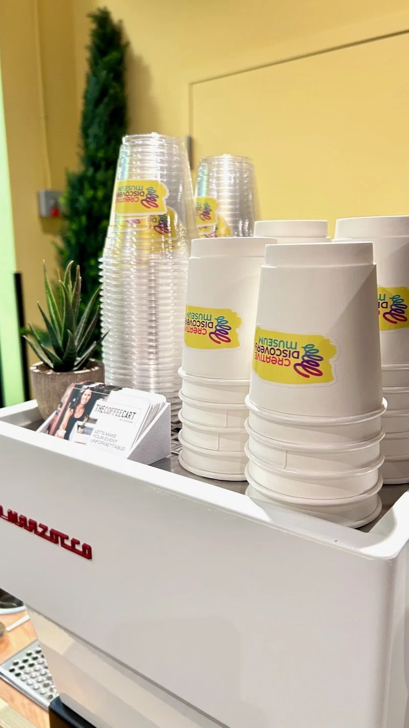 Stacked disposable cups with custom logo, a small potted succulent, and clear plastic cups on a coffee machine tray.