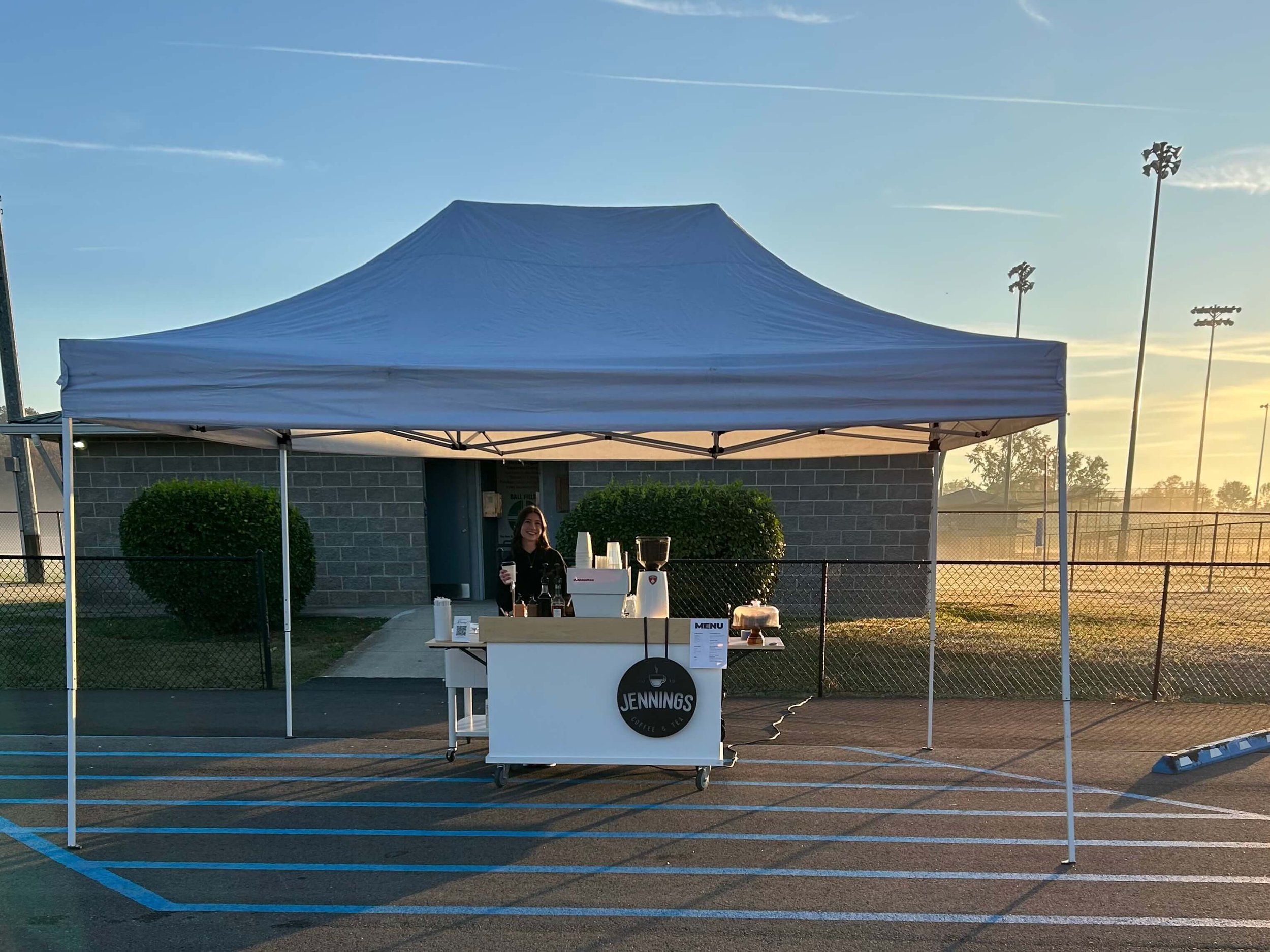 A woman standing behind a coffee stand under a white canopy tent in a parking lot at sunrise, with tennis courts and stadium lights in the background.