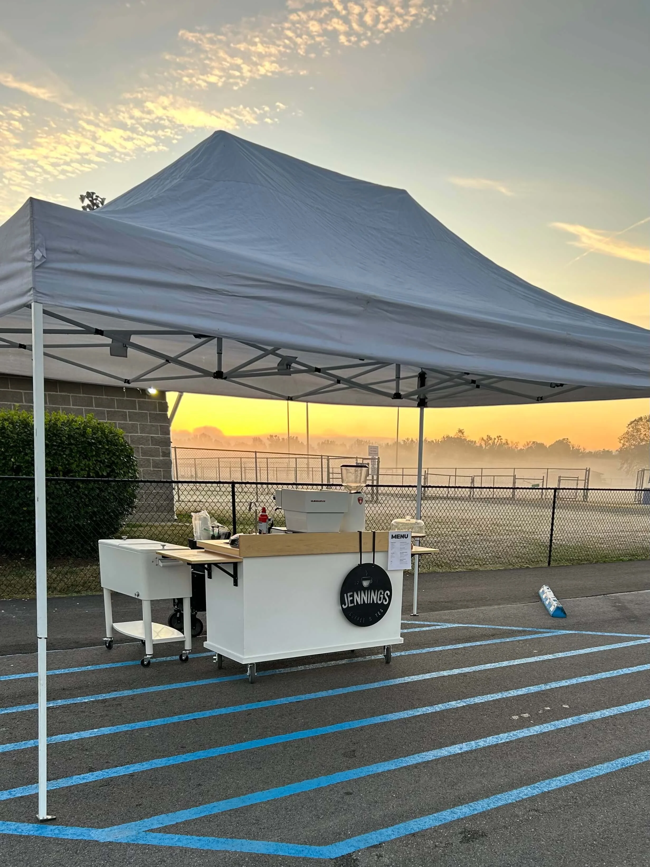 A mobile coffee stand under a white canopy tent on a parking lot at sunrise, with a foggy background and a tennis court behind a chain-link fence.