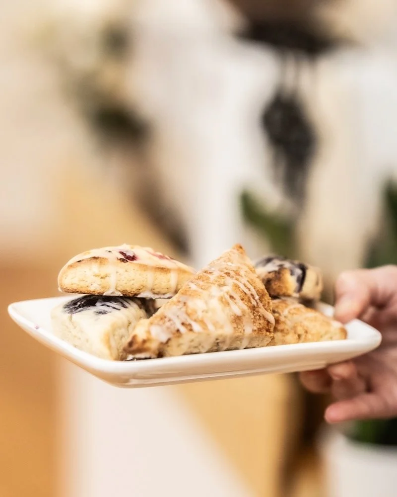 A white plate with assorted scones, including blueberry and cinnamon varieties, being held by a person. The background is blurred with neutral tones.