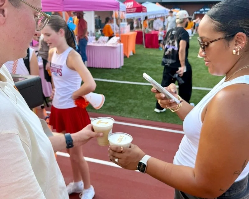 Two women at an outdoor event exchanging drinks and looking at a phone, with colorful tents and other people in the background.