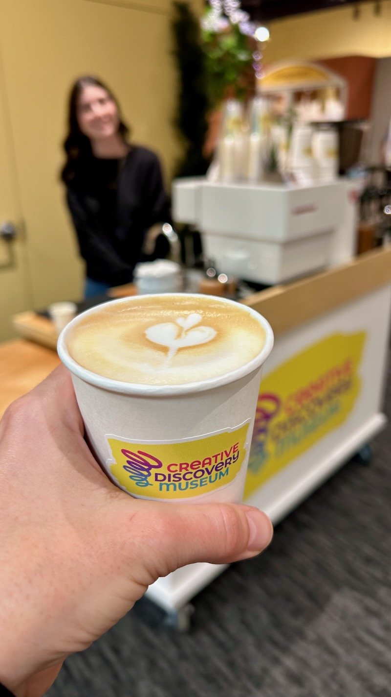 A hand holding a branded cup of latte with latte art, in front of a custom branded coffee cart with a smiling woman in a black jacket behind it.