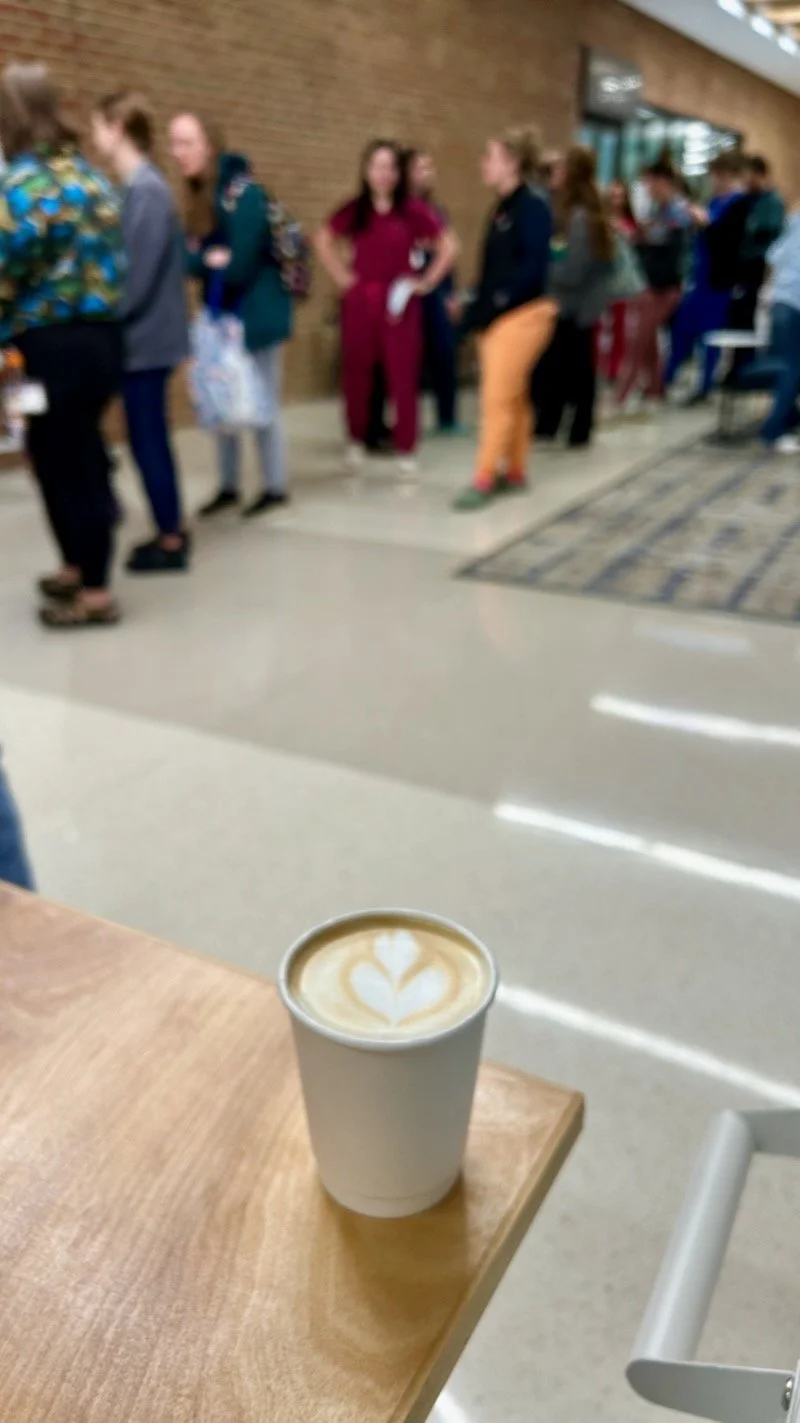 A cup of coffee with latte art sits on a mobile coffee bar in the foreground, with a blurred background of people standing in line at the College of Veterinary Medicine at the University of Tennessee Institute of Agriculture to get their coffee. 