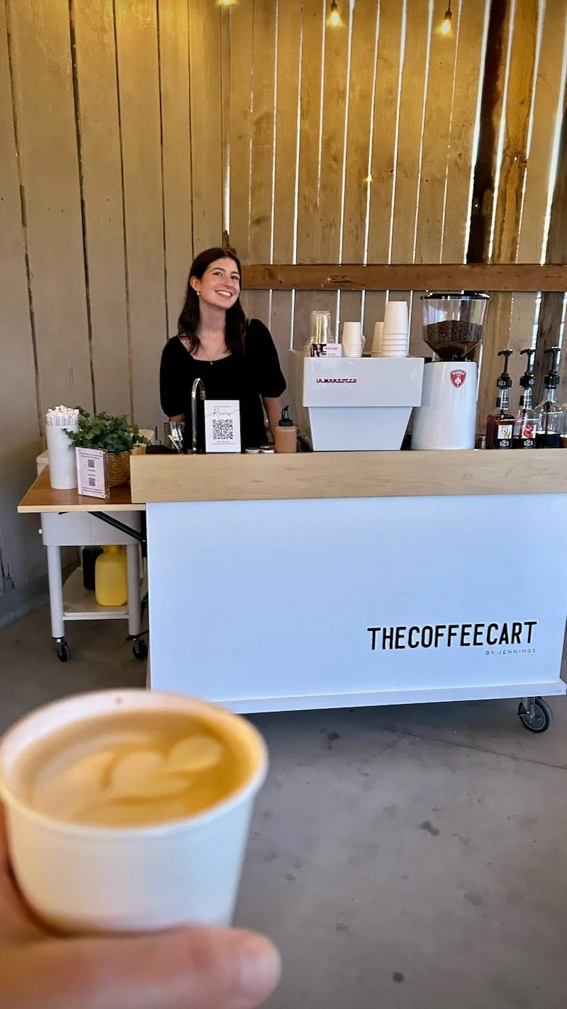 A person holding a cup of coffee in front of a coffee cart named 'The Coffee Cart' by Jennings, with a smiling barista behind the counter, in a wooden interior.