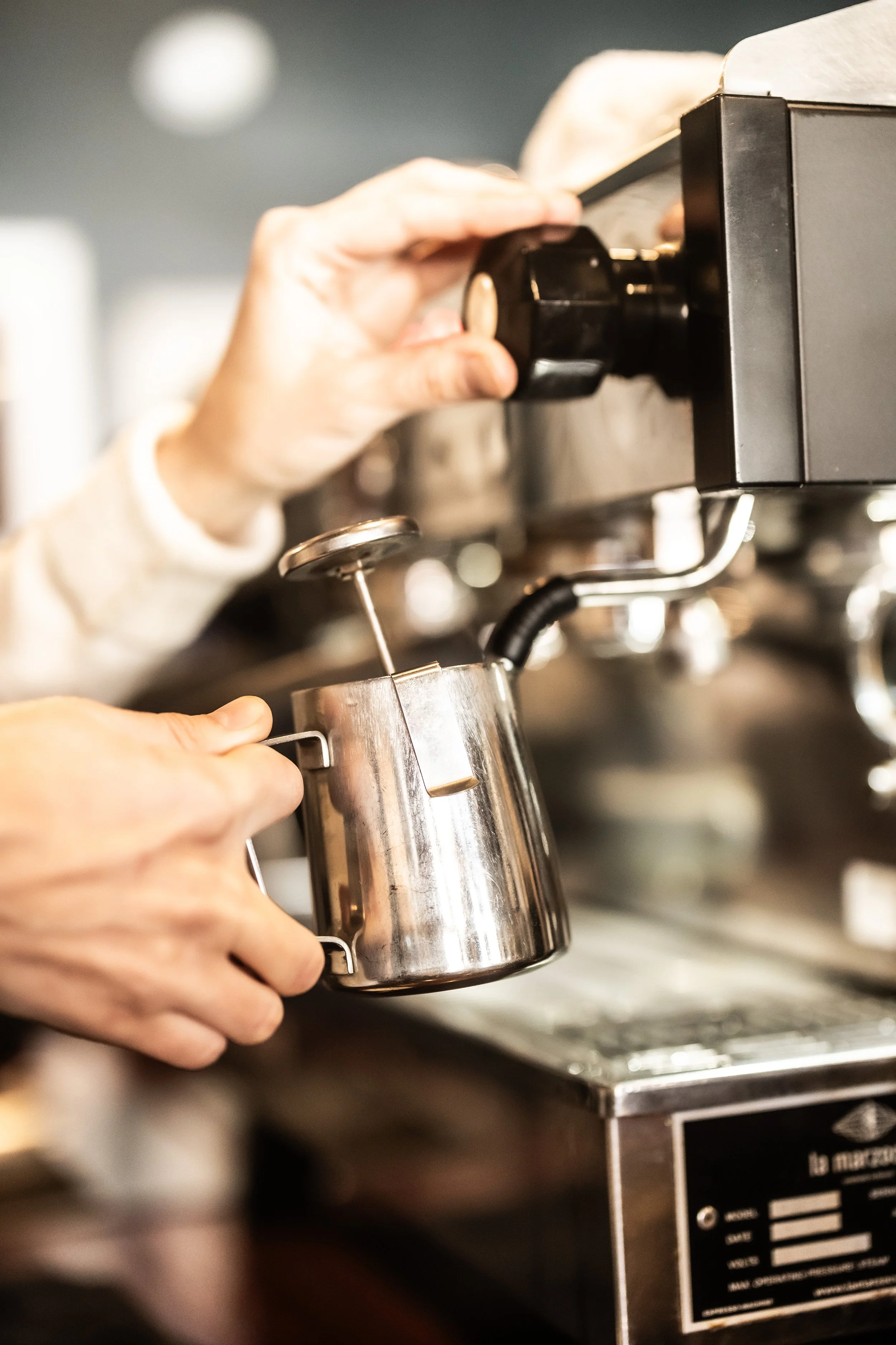 A person adjusting a coffee espresso machine, holding a small stainless steel pitcher.