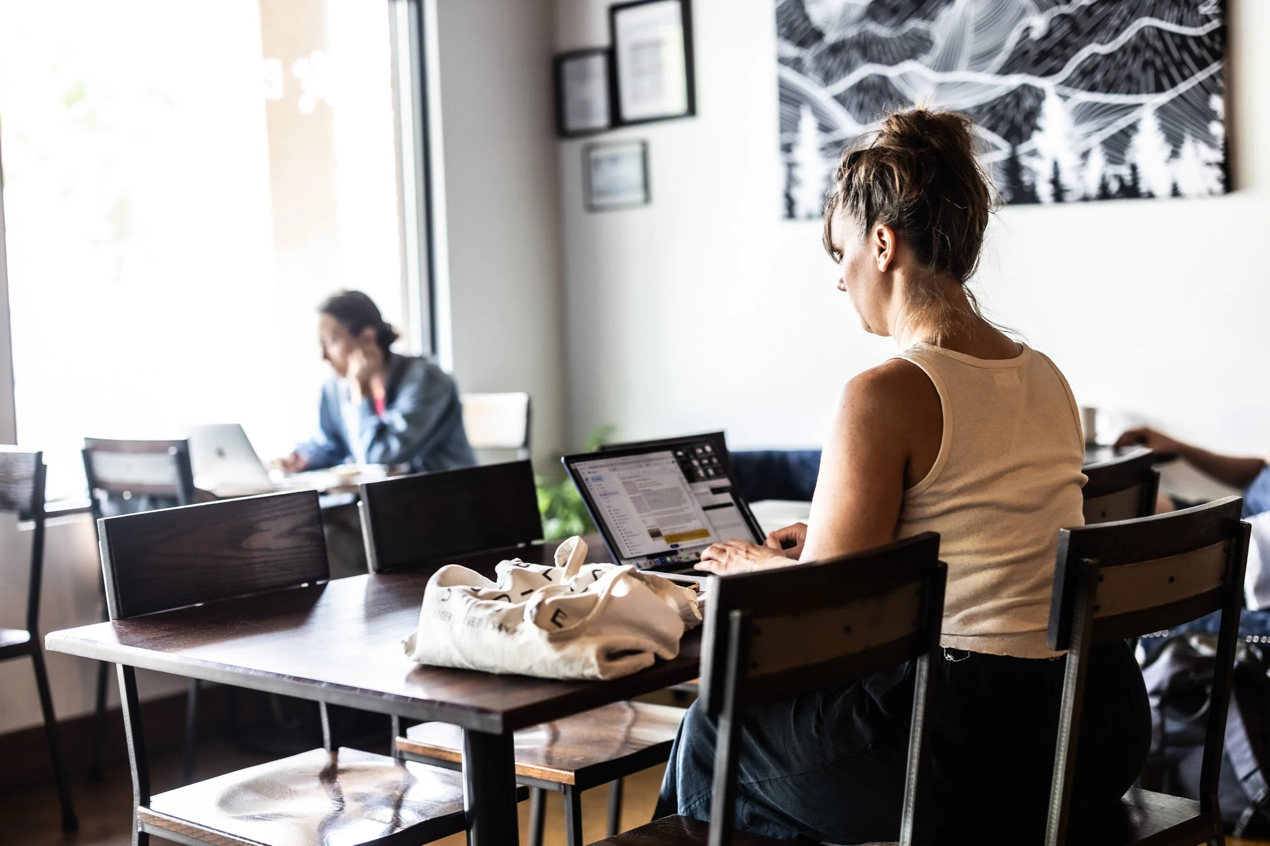 Woman sitting at a table in a cafe, working on a laptop with a tote bag next to her, other customers in the background, and artwork on the wall.