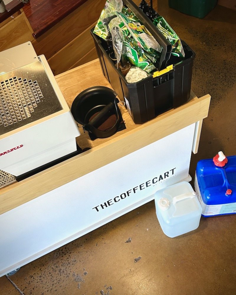 A coffee cart with coffee beans and accessories on top, a black plastic container filled with snack bags, a small black bowl, and cleaning supplies beside the cart. The cart has "THECOFFEECART" printed on the front.