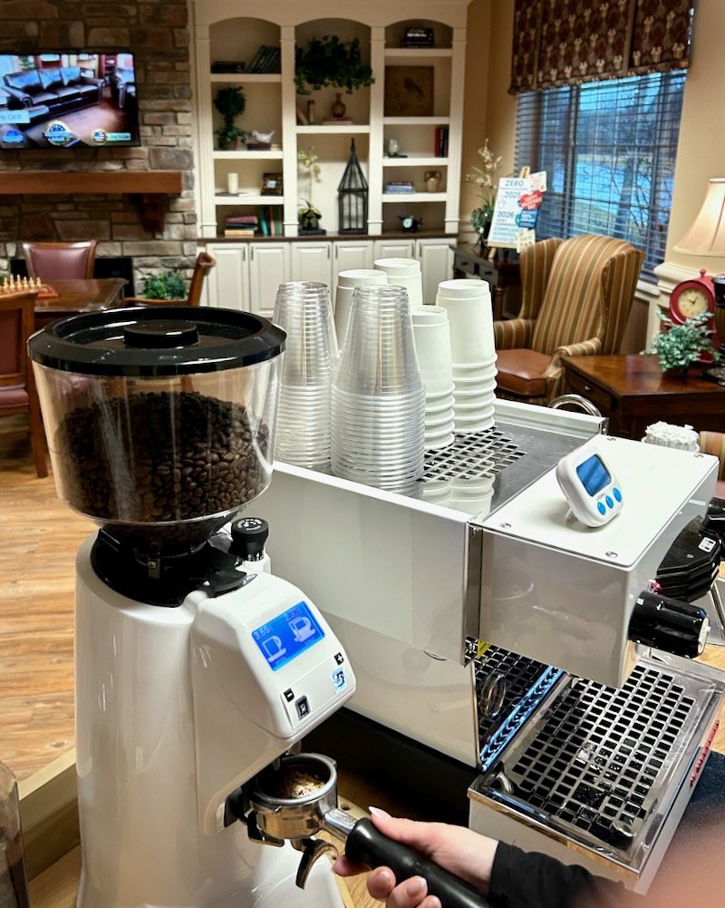 Coffee brewing setup with a coffee grinder filled with coffee beans, cups, and a barista steaming milk, in a cozy indoor setting with comfortable chairs and a large window.