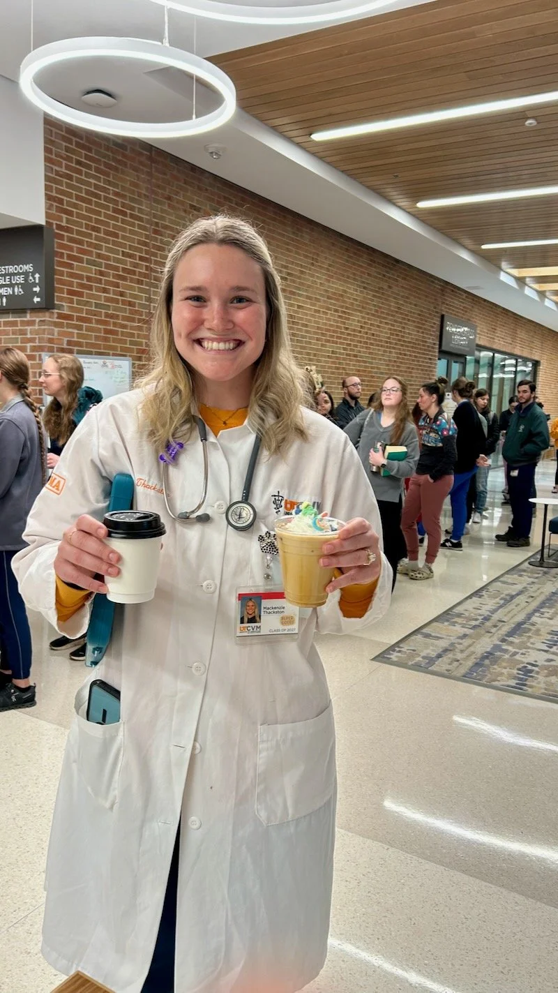 A smiling woman in a medical coat at the College of Veterinary Medicine at the University of Tennessee Institute of Agriculture holding coffes  with colorful toppings in a busy indoor setting. Smiling after getting her custom order of coffee. 
