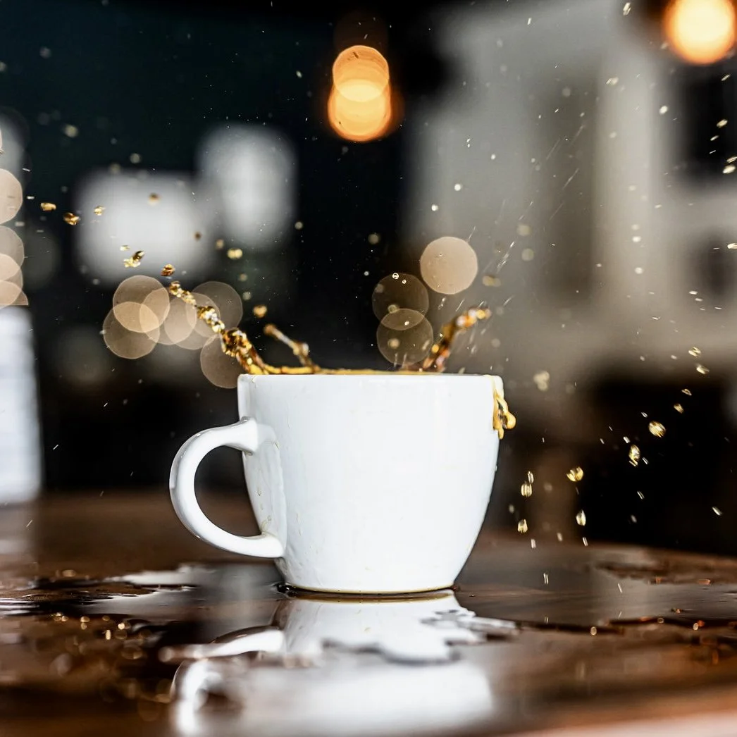 A white coffee mug with coffee spilling over the edge, creating splashes and droplets on a wooden surface with blurred city lights in the background at night.