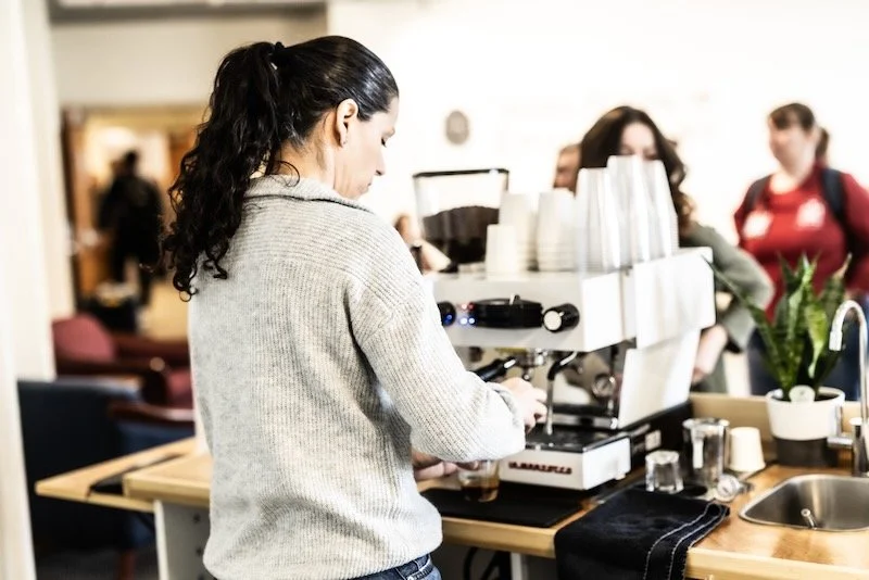 Barista making coffee behind a counter with coffee cups, and a coffee machine, while customers wait in line.