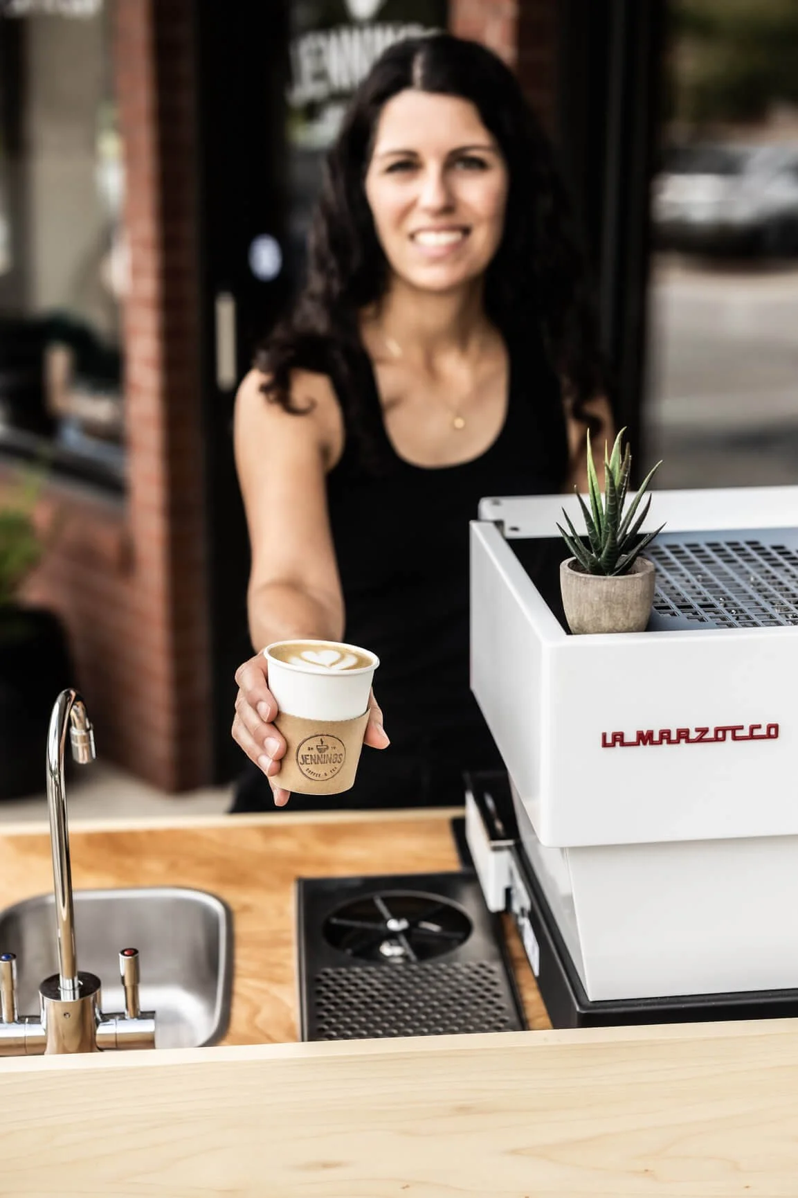 A woman with dark curly hair, wearing a black sleeveless top, is smiling and handing over a cup of coffee with latte art to the camera.