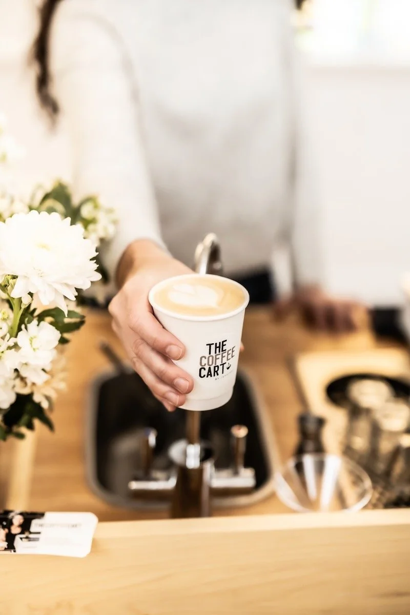 Person holding a cup of coffee at a coffee shop, with a flower arrangement on the counter and barista tools in the background.