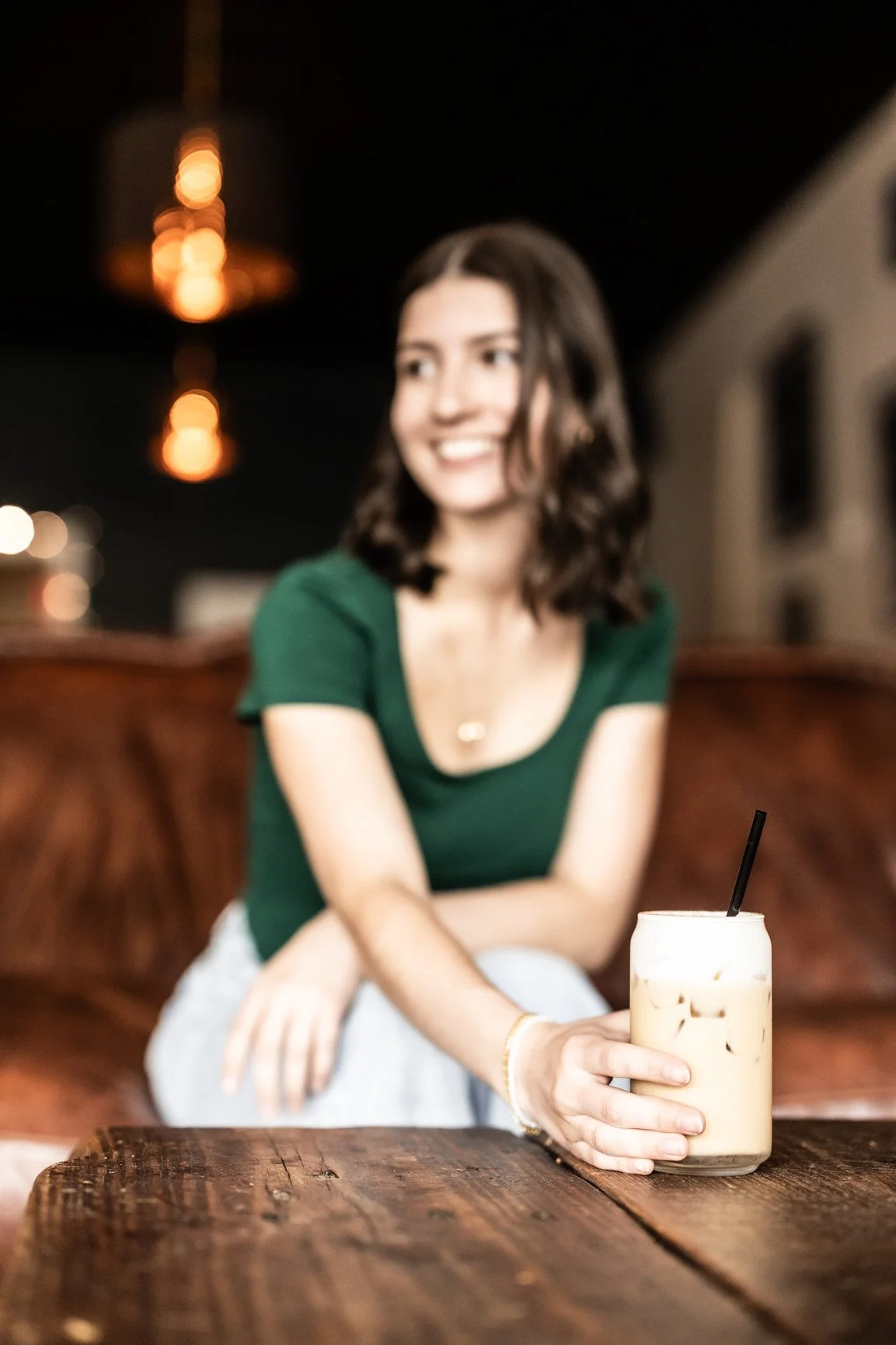 A woman with dark wavy hair in a green shirt sitting at a wooden table in a dimly lit cafe, holding an iced drink with a straw, smiling and looking to the side.