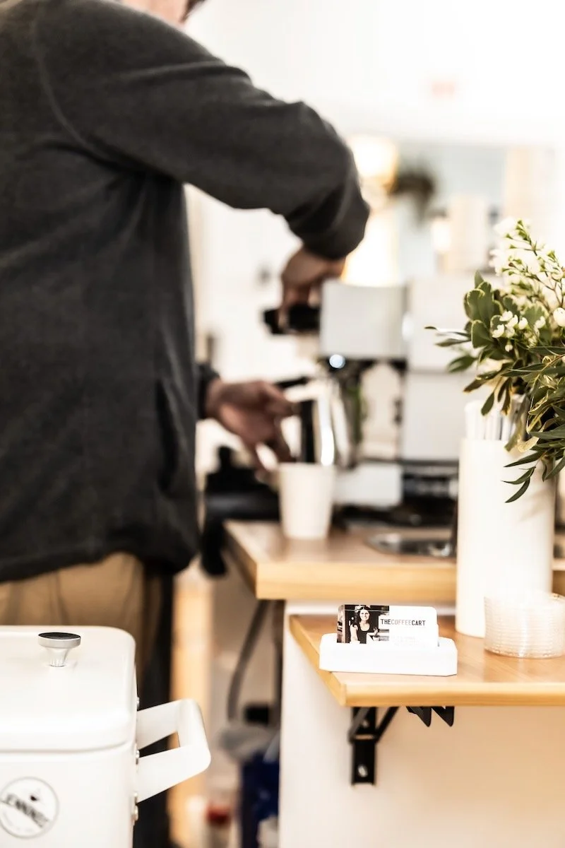 Barista preparing coffee behind the counter at a coffee shop, with a focus on a nearby vase of flowers and business cards on the counter.