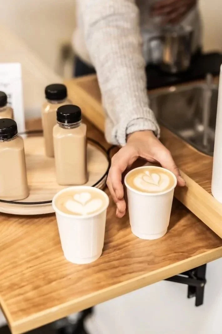 A person serving two cups of coffee with latte art on top in a coffee shop. The coffee cups are white and placed on a wooden surface, with bottles of syrup or cream nearby.