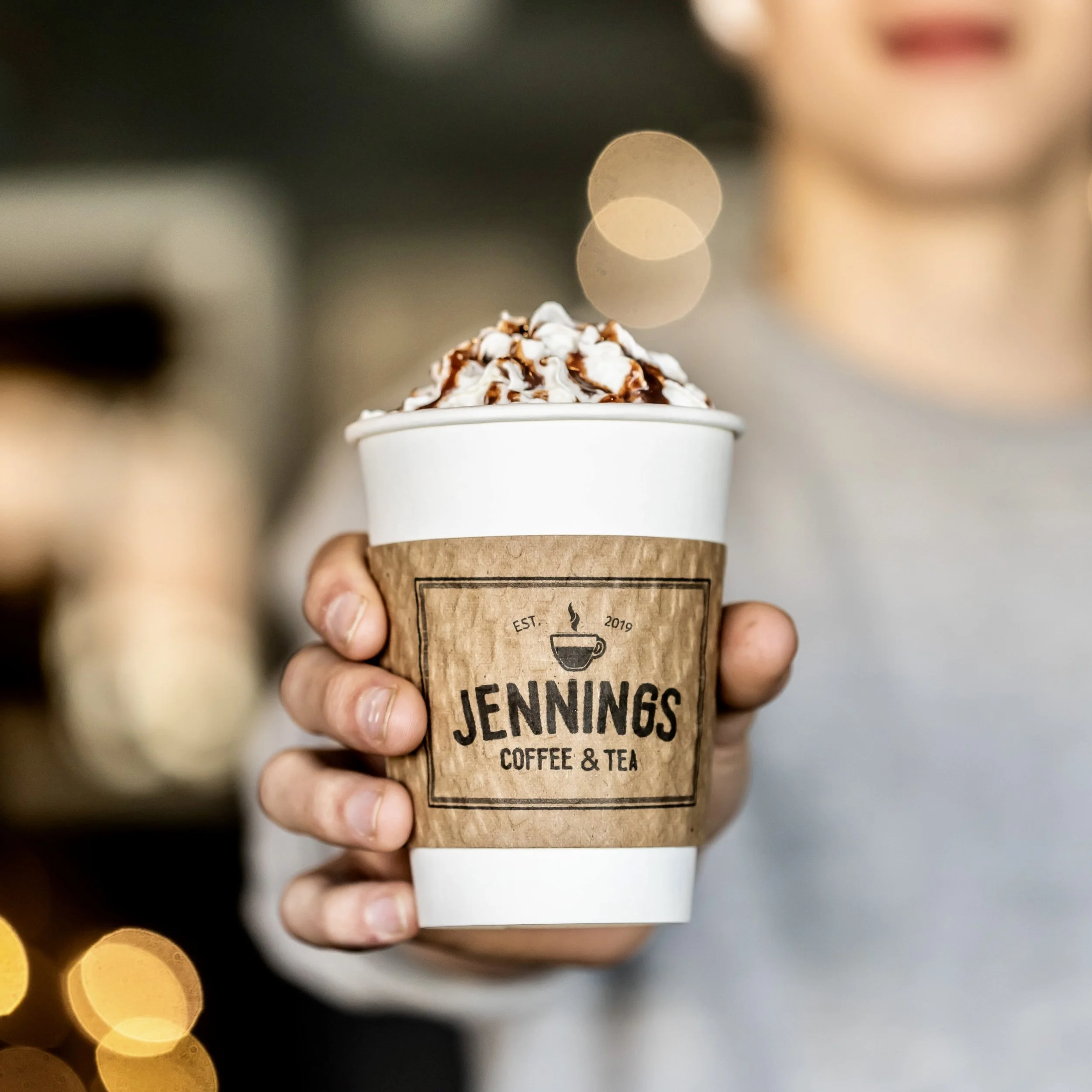 A person holding a coffee cup with whipped cream and chocolate drizzle, with a blurred background and bokeh lights.