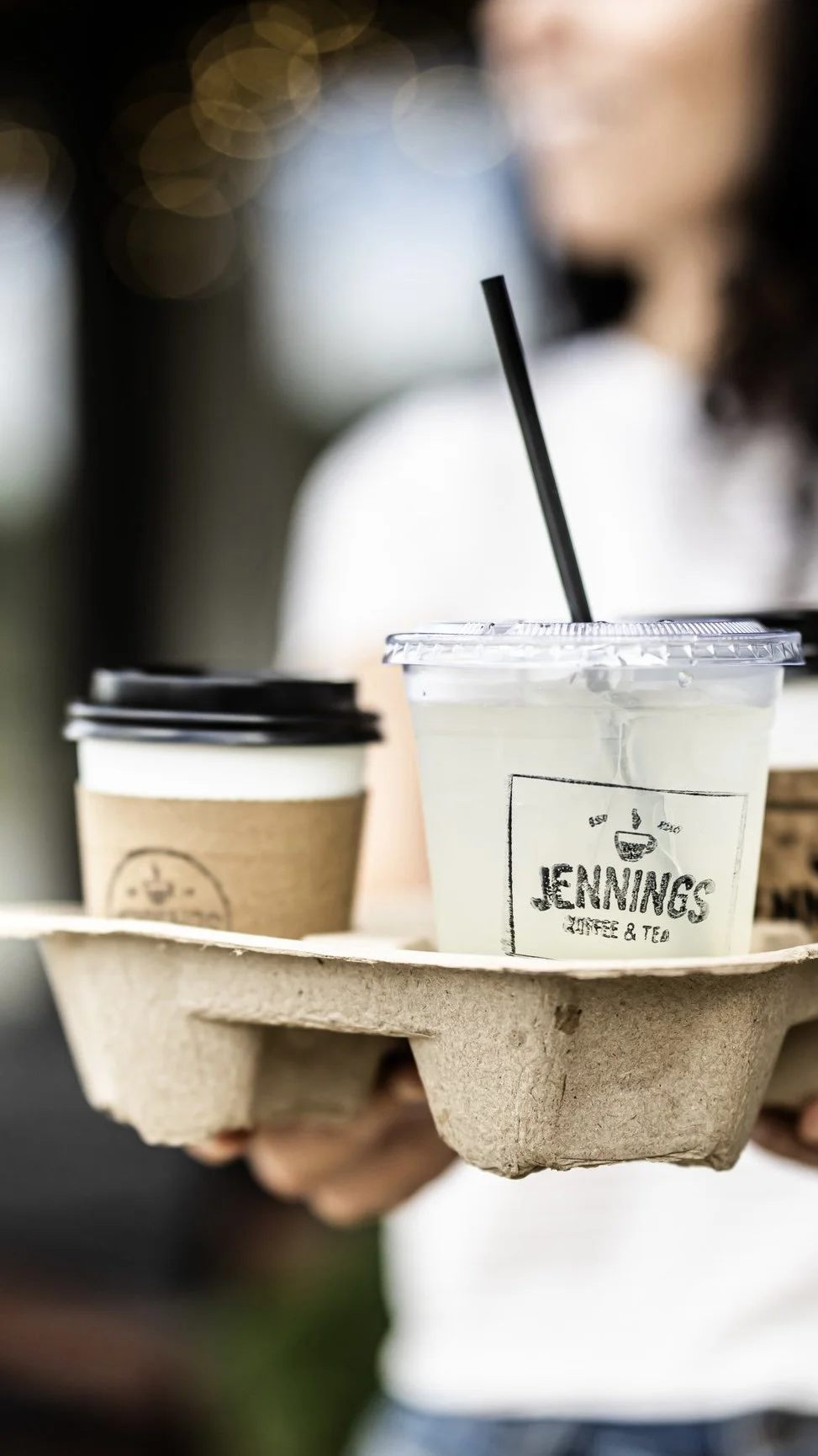 A person holding a cardboard tray with a clear plastic cup of lemonade with a black straw and a white paper cup with a black lid, both from Jennings Coffee & Tea.