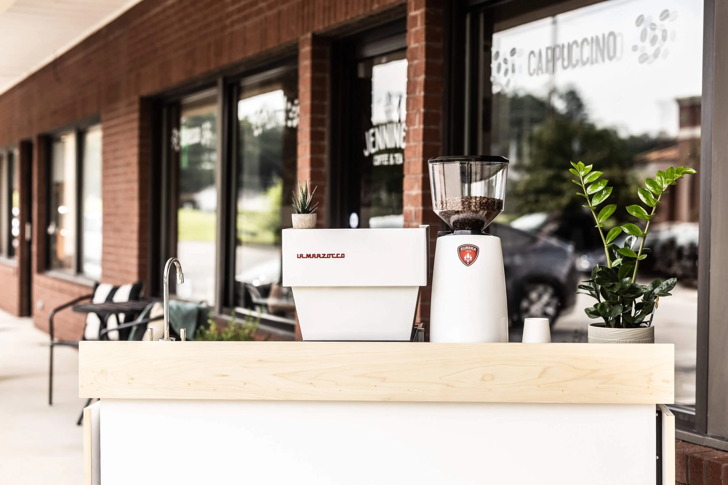 Outdoor coffee stand with a coffee grinder, a potted plant, and a small succulent in front of a brick building with large windows.